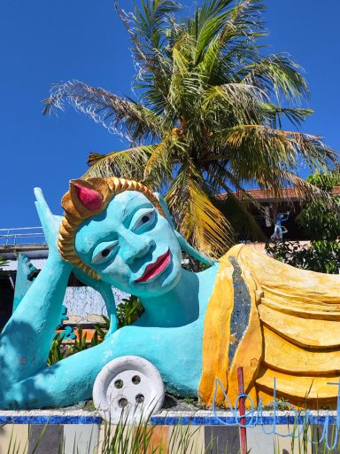 Symon's Art Zoo Bunte Statue einer liegenden Figur mit Palmen im Hintergrund.